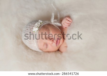 A portrait of a beautiful seven day old newborn baby girl wearing a white, knitted, mohair bonnet and rose headband. She is sleeping in a bed of cream colored wool batting.
