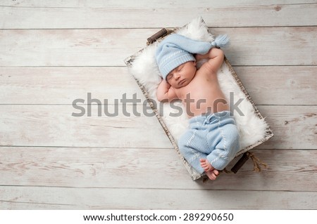 Newborn baby sleeping in a wooden crate on a whitewashed wooden floor. He is wearing light blue, upcycled pajamas with a matching sleeping cap.