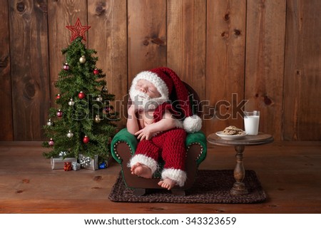 Two week old, newborn, baby boy wearing a crocheted Santa suit with beard. He's sleeping on an armchair. Shot in the studio with props, such as a Christmas tree, glass of milk and crocheted cookies.