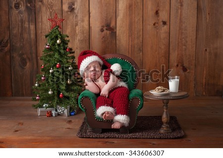 Two week old, newborn, baby boy wearing a crocheted Santa suit. He is sleeping on a tiny armchair. Shot in the studio with props, including a Christmas tree, glass of milk and crocheted cookies.