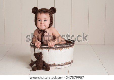 Nine month old newborn baby girl wearing a brown, crocheted bear hat. She is sitting in a white, wooden bucket and has a stuffed bear toy. Shot in the studio on a white, wood paneled background.