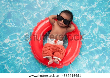 Two week old newborn baby boy sleeping on a tiny inflatable swim ring. He is wearing crocheted board shorts and black sunglasses.