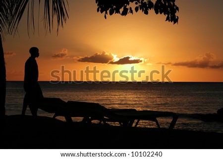 a sunset in Barbados with the silhouette of a man and two sunbeds, focussed on the man with slightly de-focused clouds