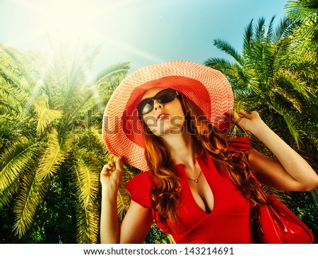 Young beautiful woman in red fashion dress, big hat and sun glasses on tropical resort