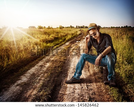 Young handsome man traveler sitting on his suitcase on a dirt road outdoor