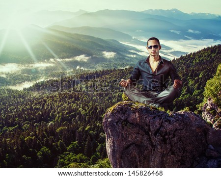 Young fashion man in dark blue shirt and gray pants sitting isolated on mountains stone