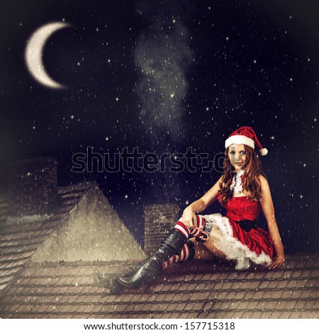 beautiful sexy christmas fairy woman  in red dress and in santa hat sitting on tiled roof of house at magic night