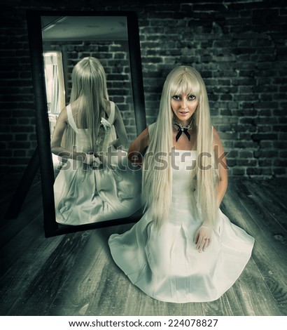 Young beautiful blond woman sitting on wooden floor in old dark room with big knife in mirror reflection