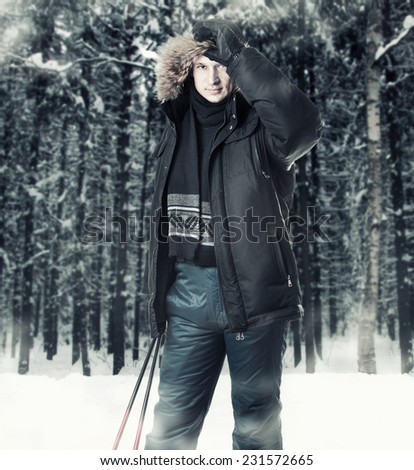 Young man skier wearing black fur hood winter jacket and holding sticks in winter forest