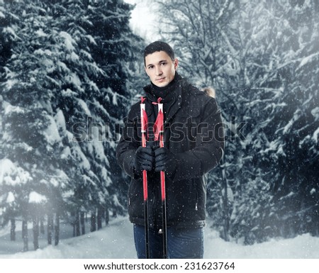 Young handsome man skier wearing black fur hood winter jacket Alaska and pants holding sticks in snowy forest