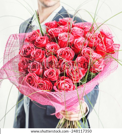 Valentines day or proposal. Young happy handsome man holding big bunch of red roses in his hand on grey background, studio shot