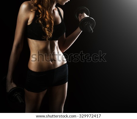 Dark contrast photo of young beautiful fitness woman with beads of sweat which training in gym. Working out with dumbbells on black background