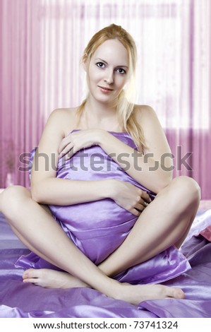 Seductive young woman sit on silk sheet holding pillow on bed in morning and smiling