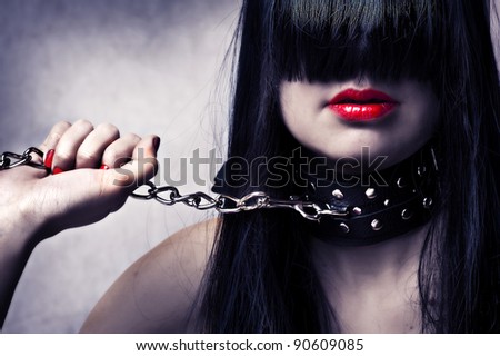 Fashion portrait of young beautiful female model. Glamour woman with long black hair and sexy hairstyle. Lady with leather collar with studs on a metal chain in hand. Red make-up on lips and manicure