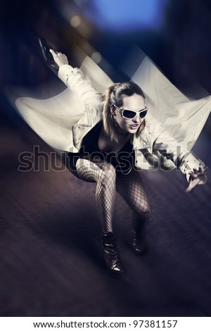Portrait of beautiful woman with gun in white raincoat and designer sun glasses jumping on night  street. Motion