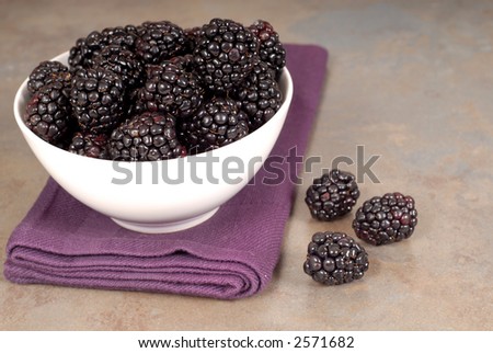 Sweet blackberries in a white bowl on a purple napkin with blackberries on the side