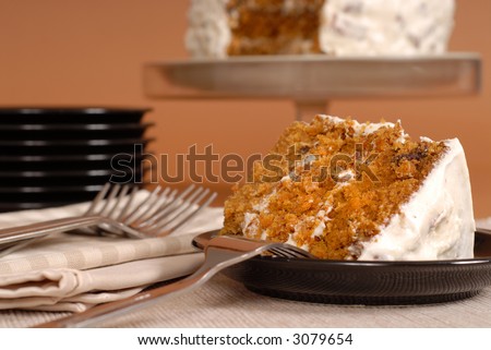 A piece of carrot cake with forks, plates, and whole cake in background