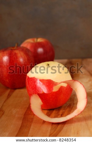 A partially pealed apple on a cutting board with two apples in the background