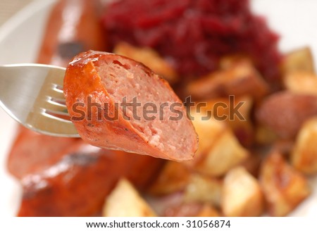Piece of freshly grilled smoked sausage on a fork with potatoes and red cabbage