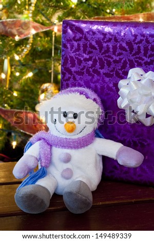 Smiling stuffed snowman with a purple present and a Christmas tree