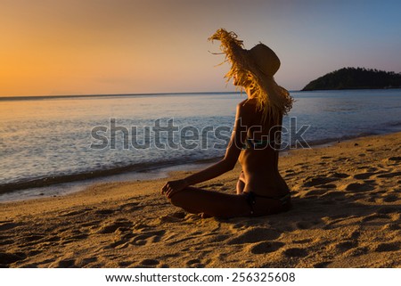Slim woman in big hat is sitting on the beach and looking to the sunset