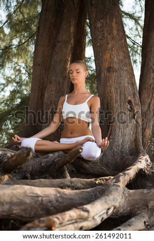 Pretty slim Woman in white costume doing Yoga pose near big tree
