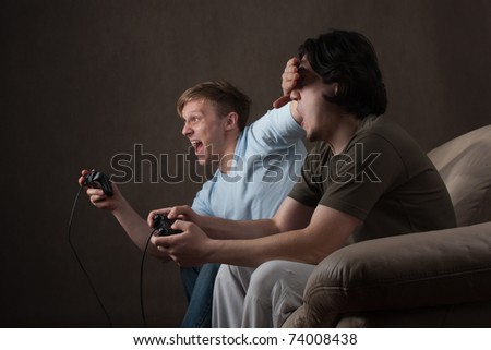 young guy covering friend\'s eyes while playing video games on gray background