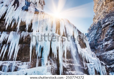 Icicles and a jet of water on the frozen waterfall. Beautiful winter landscape with mountain gorge at sunset