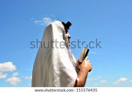 A Jewish man wearing Tallit and Tefillin read from the Torah book pray to God under a blue sky with sheep clouds on Jewish holiday.
