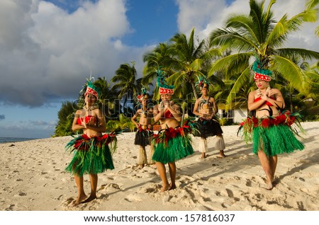 Group portrait of Polynesian Pacific Island Tahitian dance group in colorful costumes dancing on tropical beach with palm trees in the background.