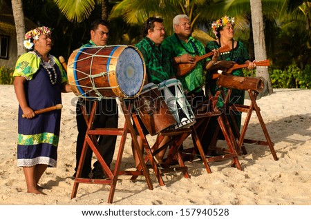 Group portrait of Polynesian Pacific Islanders band plays Tahitian music on tropical beach with palm trees in the background.