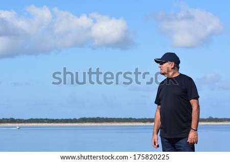 A color portrait photo of a fit mature man in his 70\'s wearing a blue t\'shirt and a baseball cap against a blue sky with white clouds background.
