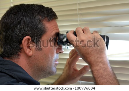Man (age 35-40 ) looks and searches with binoculars and  looks out through Venetian blinds. Concept photo of curious, spy, nosy man.