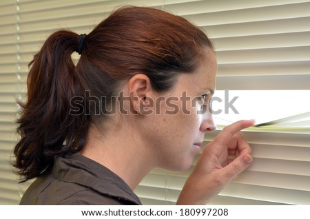 Young woman (age 25-30 ) looks out through Venetian blinds. Concept photo of curious, spy, nosy woman.