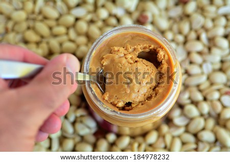 Man hand holds a spoon with crunchy peanut butter in jar with dry roasted peanuts in the background. Concept photo of food