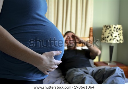 Pregnant woman holds her abdomen and distress relaxed man lying on double bed in the background. Concept photo of pregnancy, pregnant woman lifestyle. copyspace