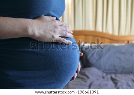 Pregnant woman holds her abdomen with double bed in a  bedroom in the background. Concept photo of pregnancy, pregnant woman sex life. copyspace