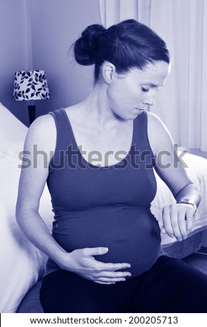 Young pregnant woman looking at her wrist watch, timing contraction pains and holding her tummy in her bedroom at home. (BW)
