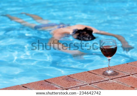 A cup of red wine on a pool side with drunk drowning person (woman) in a swimming pool at the background. Concept photo