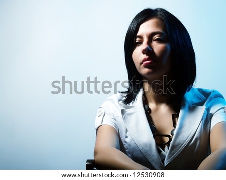 A high-key portrait about a pretty trendy woman with black hair who is lighted in blue, she is sitting on a chair and she has a charming look. She is wearing a white coat and a stylish necklace.