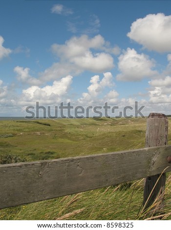 View of grassy dunes by the North Sea in Northern Holland with country fence in the foreground, bright blue sky, and some figures walking in the distance to give perspective to the landscape.