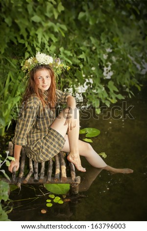 A girl in wreath sitting on the bank of a river