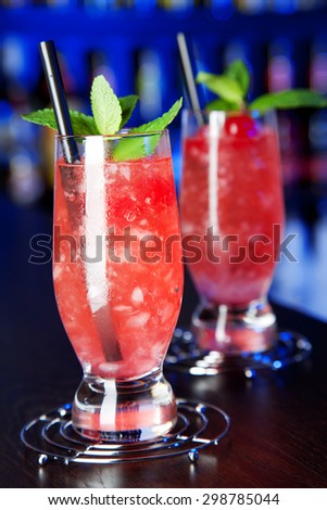 Two glasses of Eclipse cocktail at the bar, lit in blue light. Ingredients: 3 oz gin 1 oz raspeberry liqueur 1/2 grenadine crushed ice to the top mint and maraschino cherry for decoration