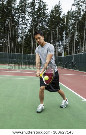 Young man hitting two handed backhand on outdoor tennis court with trees in background