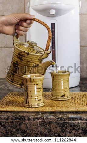 Hand pouring tea into cup on kitchen counter top with hot water dispenser in background