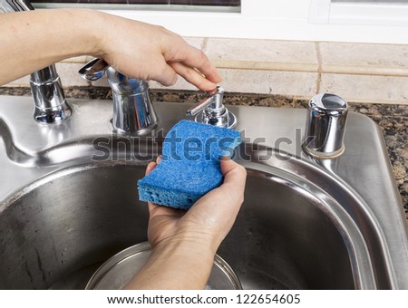 Female hand pushing soap dispenser for soap to put on cleaning sponge with stainless steel sink and pan in background