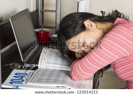 Mature Asian woman with head down on income tax tables booklet with computer, coffee  cup and glasses on desk