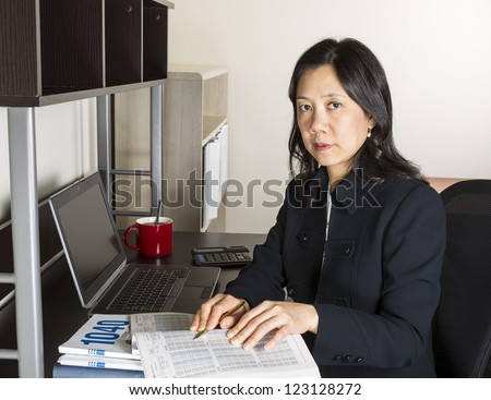 Professional Mature Asian woman working on Personal Income Taxes with tax table booklet, computer, coffee cup with spoon on desk
