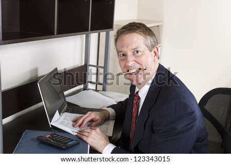 Mature man chewing pen while working on income tax with calculator, computer and papers on desk