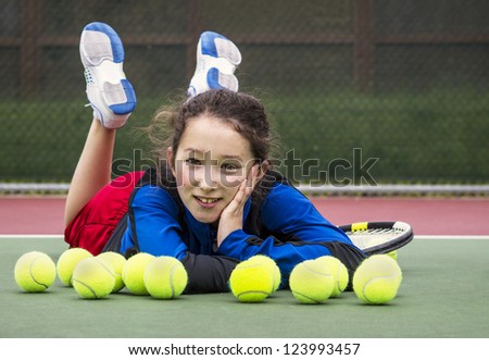 Horizontal portrait of smiling teenage girl tennis player laying on the outdoor court behind a row of tennis balls with head resting in her hand and feet in air